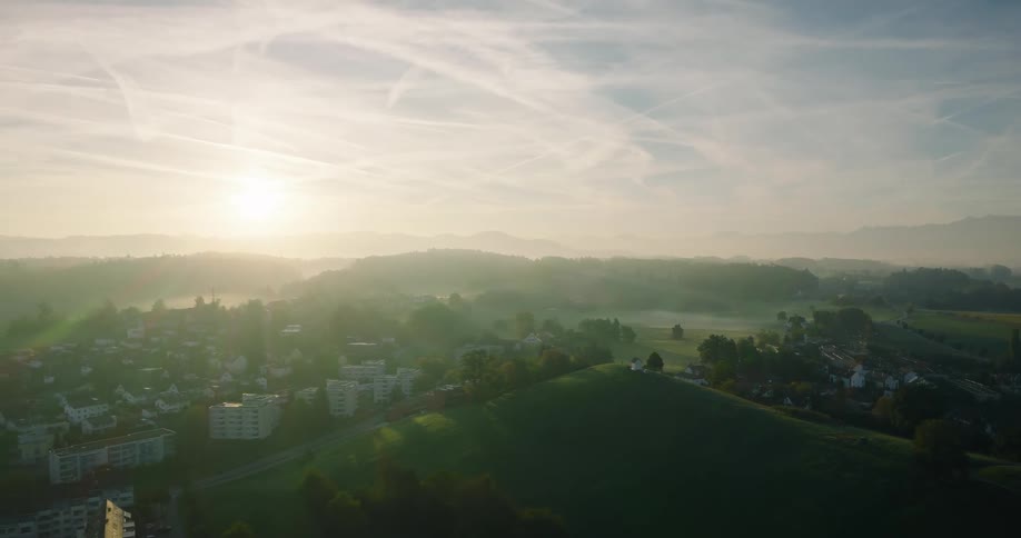 An expansive aerial shot reveals a picturesque town waking up to a misty sunrise amidst rolling green hills