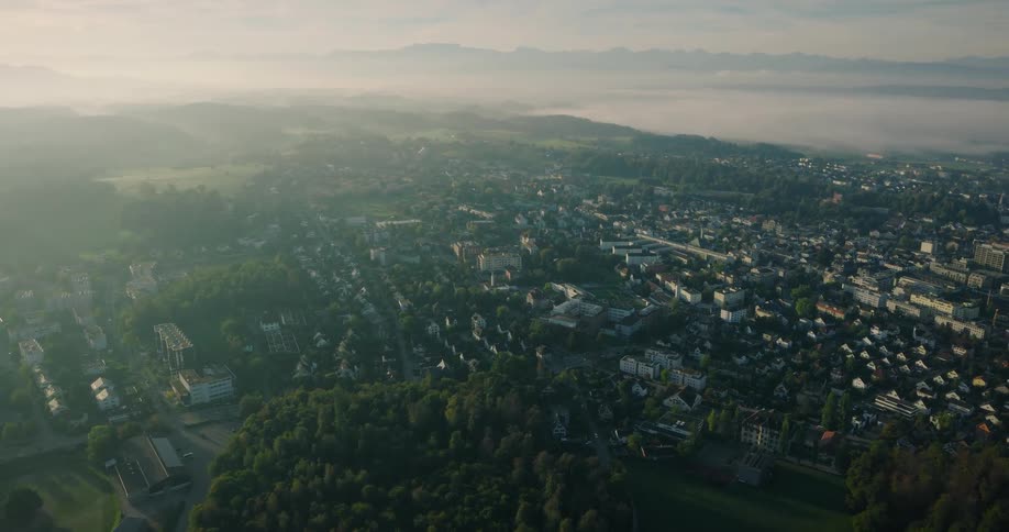 A slow aerial ascent reveals a picturesque European town surrounded by lush green forests at sunrise, with a blanket of fog in the valleys and mountains beyond