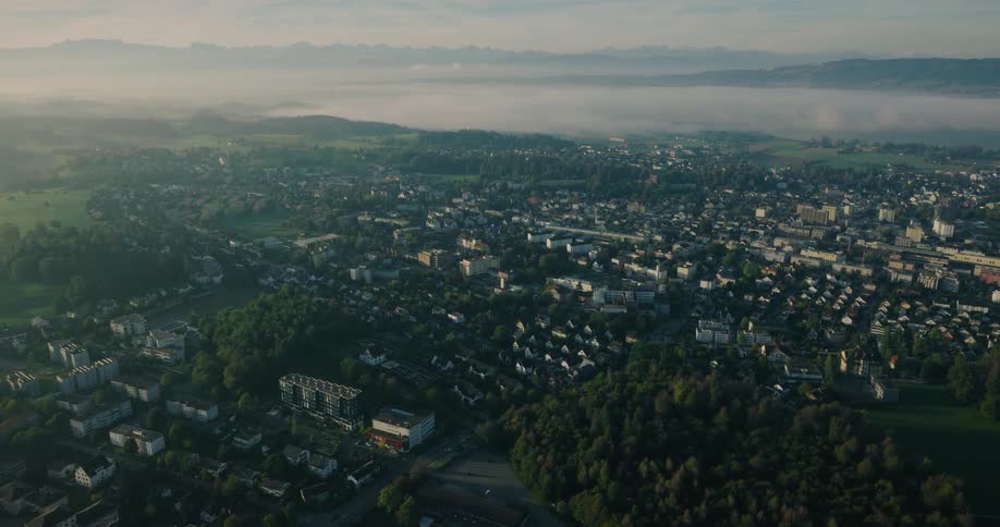 A static aerial shot captures a tranquil town nestled in a verdant valley during a foggy sunrise, with majestic mountains in the distance