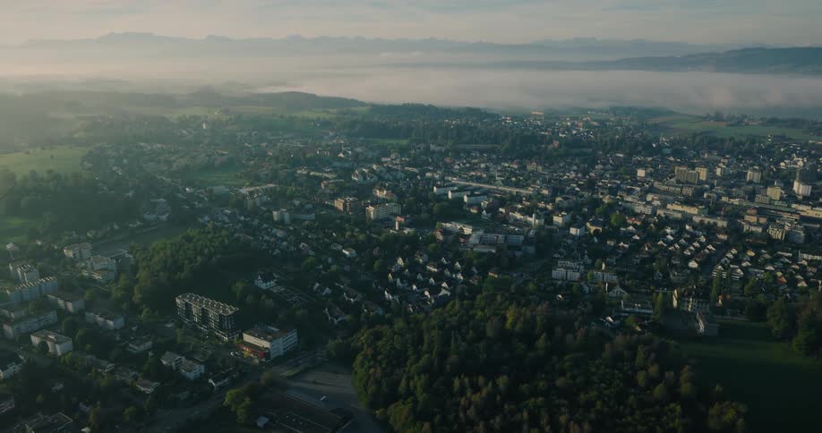 Static aerial shot of a small city in the Swiss countryside during a misty sunrise with the Alps in the background
