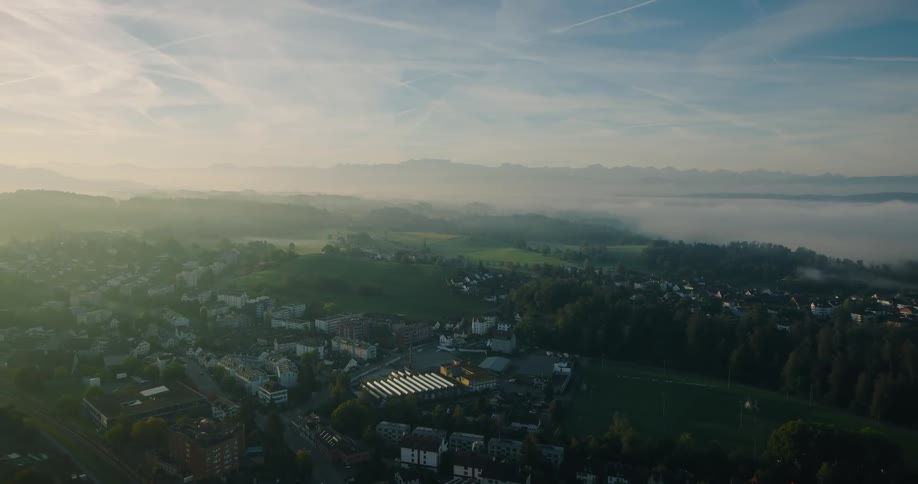 An aerial view captures a quiet European town in a misty valley at sunrise, with a majestic mountain range on the horizon