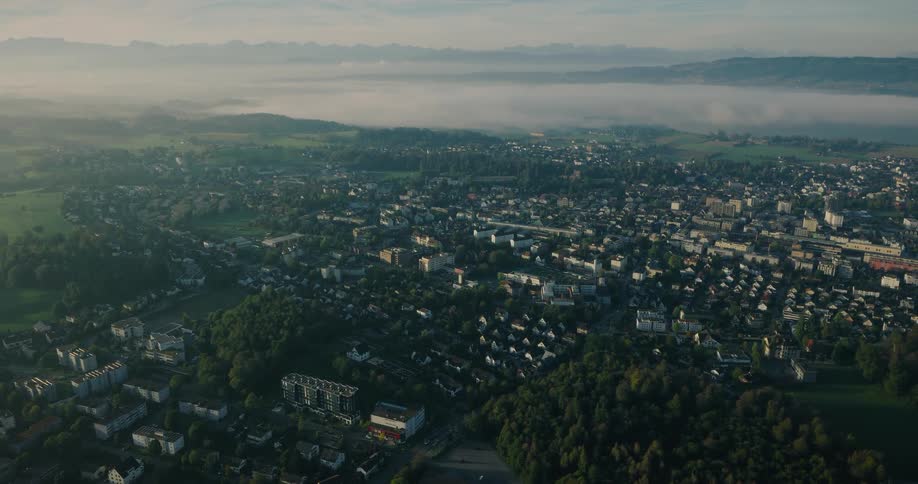 A static, wide aerial shot shows a tranquil European town nestled in a green valley, with a layer of morning fog and a mountain range on the horizon