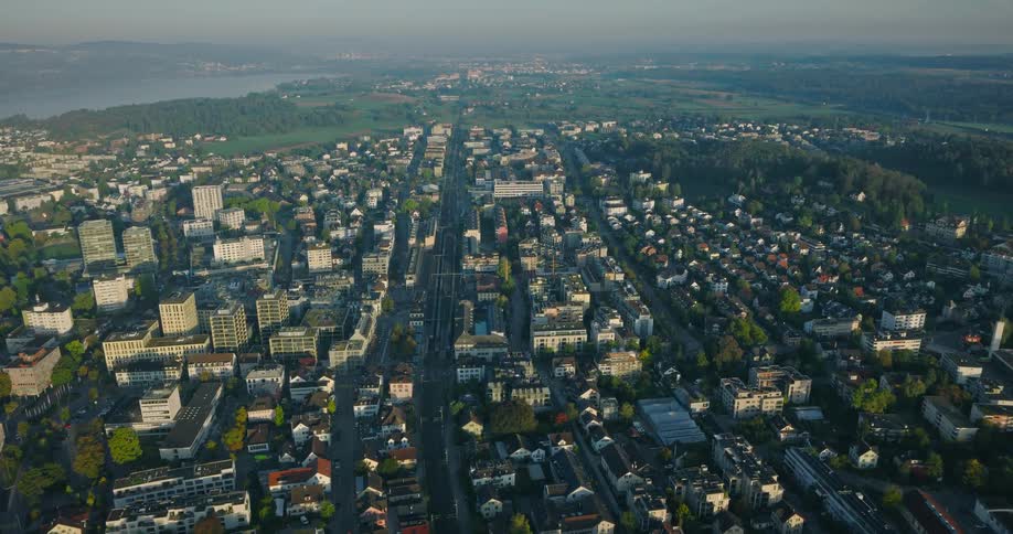 A stunning high-altitude aerial shot slowly flies over a European city during the golden morning light, with a lake and lush hills in the distance
