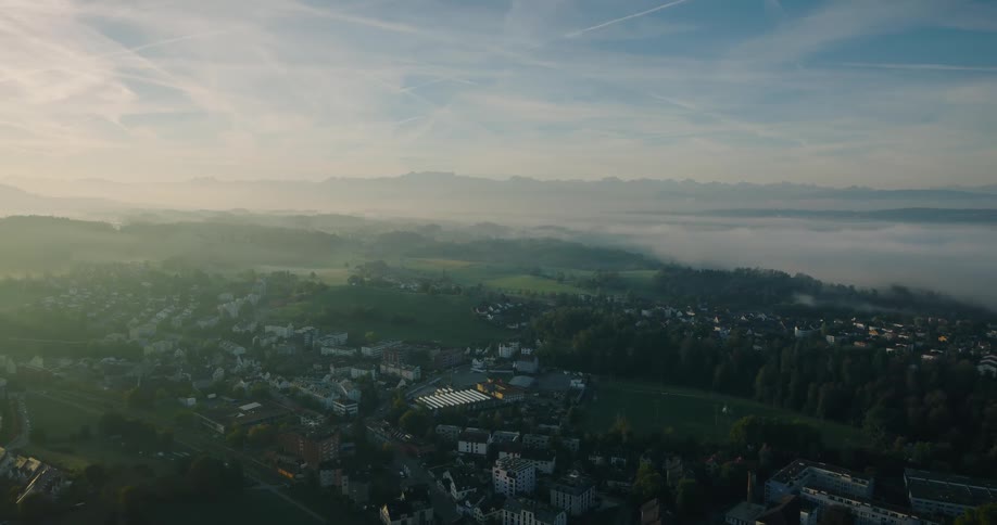 A stunning aerial timelapse captures morning fog blanketing a European town nestled in a valley, with traffic flowing and majestic mountains on the horizon