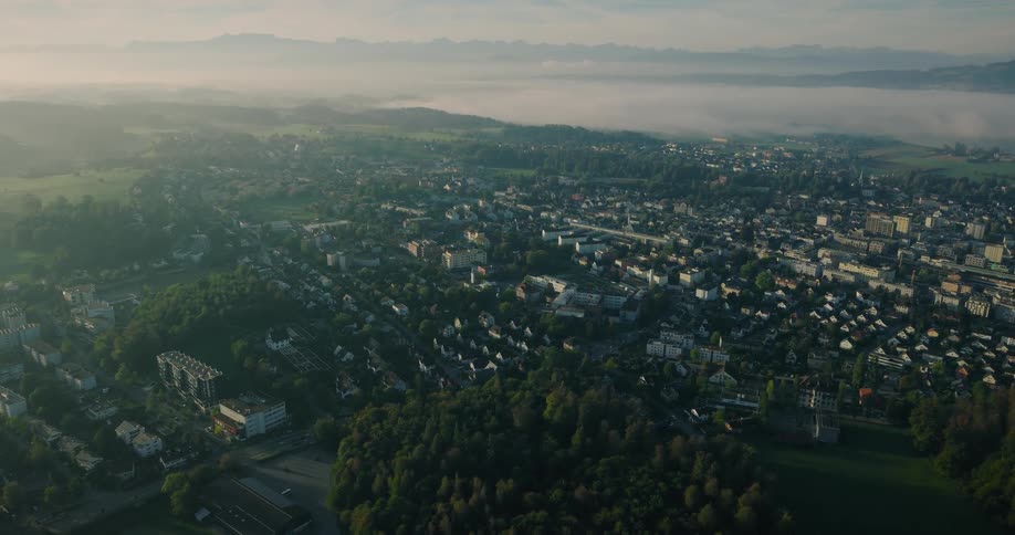A sweeping aerial view reveals a tranquil town in a green valley during a misty sunrise, with a mountain range visible on the horizon