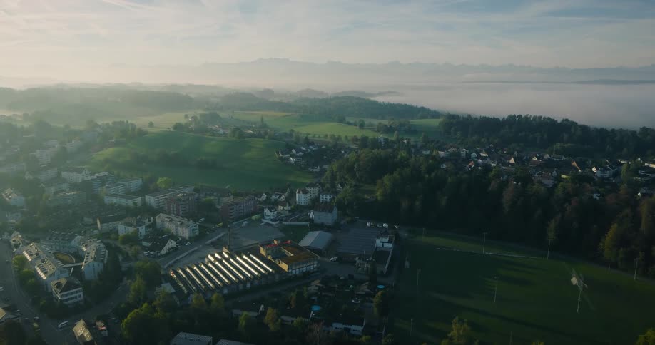 A stunning aerial view flies over a European town nestled in a lush, green valley during a beautiful foggy sunrise