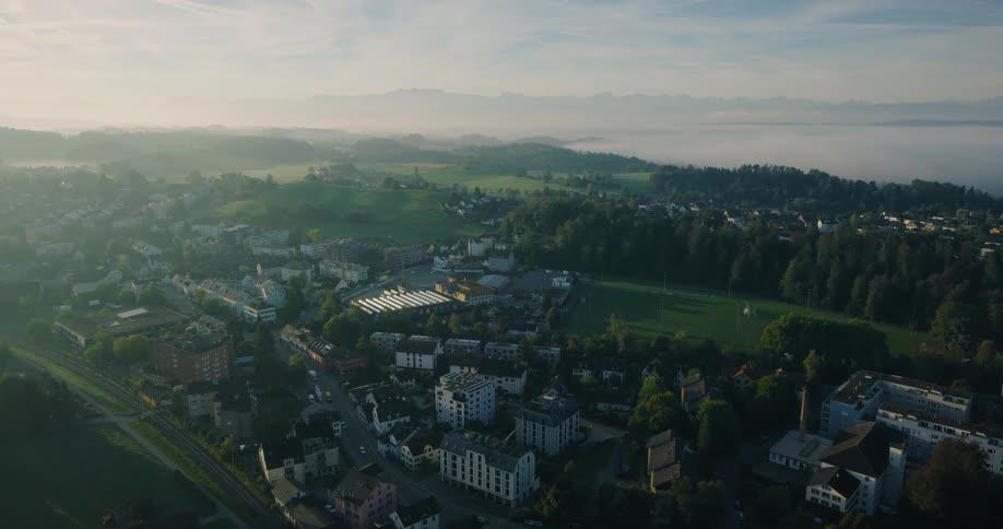 A slow aerial flyover of a charming town nestled in a green, foggy valley during a golden sunrise, with majestic mountains visible on the horizon