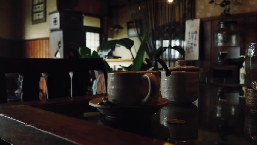 A ceramic coffee cup and sugar bowl rest on a dark, reflective table in a dimly lit, cozy cafe