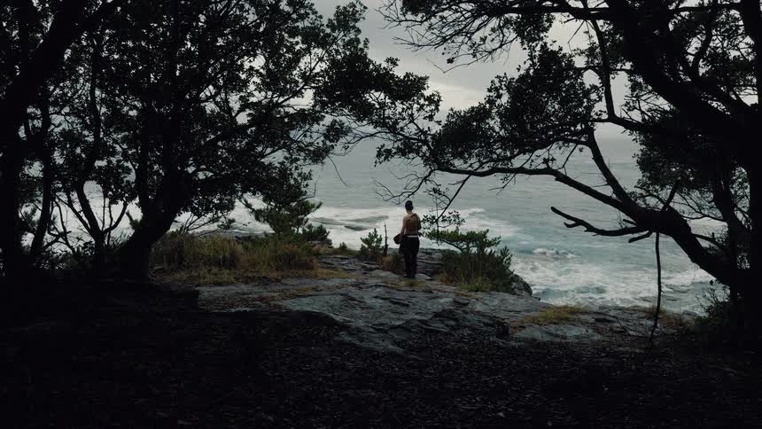 A lone shirtless man emerges from a dark forest to stand on a cliff overlooking a stormy ocean