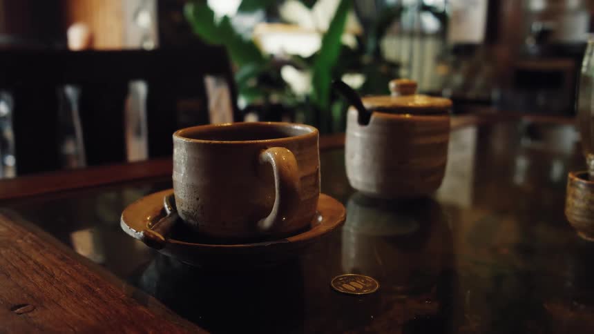 A rustic, handmade coffee cup and sugar bowl sit on a dark, reflective table in a cozy, dimly lit cafe