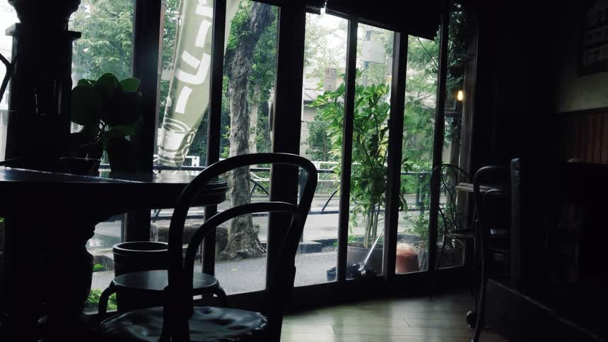 A tranquil, moody view from inside a dimly lit cafe with a silhouetted table and chairs looking out onto a rainy street as traffic passes by