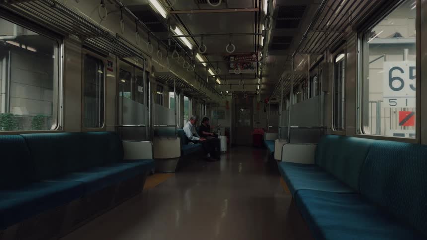 Two elderly men sit in a nearly empty Japanese train car as it travels through a town, the light flickering as it moves