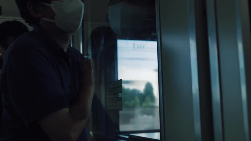 A moody shot of a passenger in a face mask standing by the door on a moving train, watching the city pass by