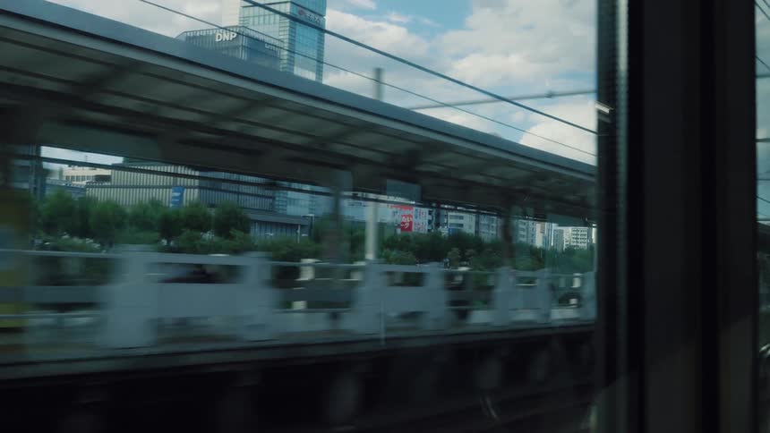 A cinematic view of the Tokyo cityscape passes by from the window of a moving train as it pulls into a station