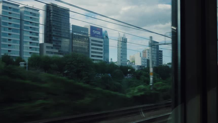 A slow motion view of the cityscape with its modern buildings and lush green trees from the window of a moving train