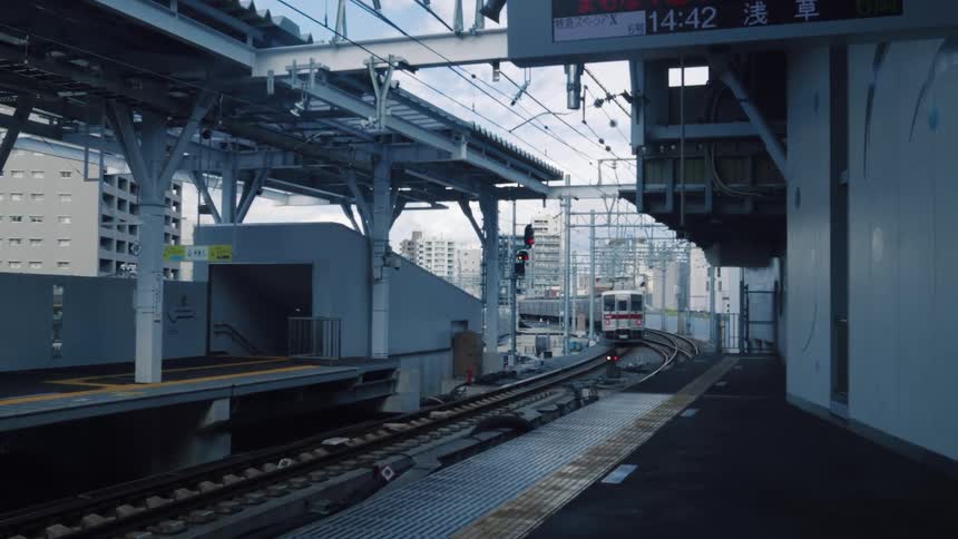 A silver commuter train departs from a modern, empty platform at a Japanese railway station during the day