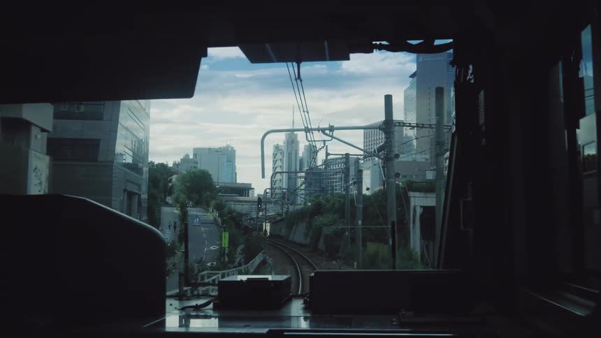 A point-of-view journey from the conductor's cabin as a train travels through a modern Japanese city, winding past skyscrapers and along complex tracks before plunging into a dark tunnel