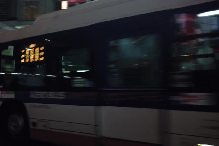 A city bus with a glowing electronic sign drives through the streets of Tokyo at night