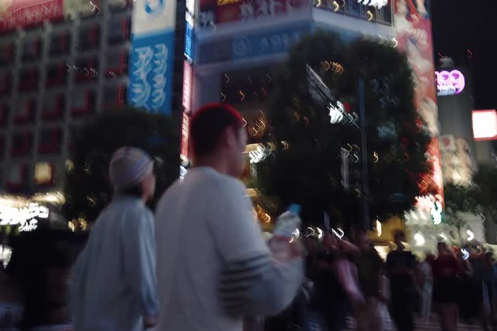 A fast-paced, shaky camera follows two young people walking through the vibrant and crowded neon-lit streets of a city at night