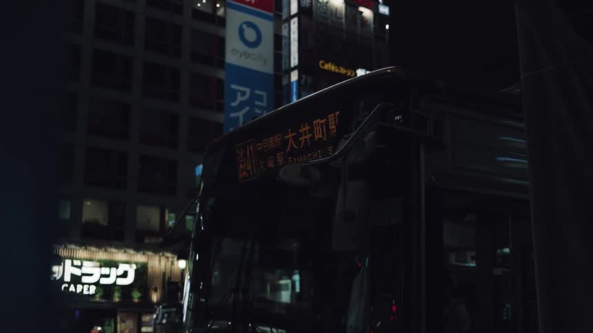 A Japanese city bus is parked at a stop at night, its destination sign glowing against the dark urban backdrop