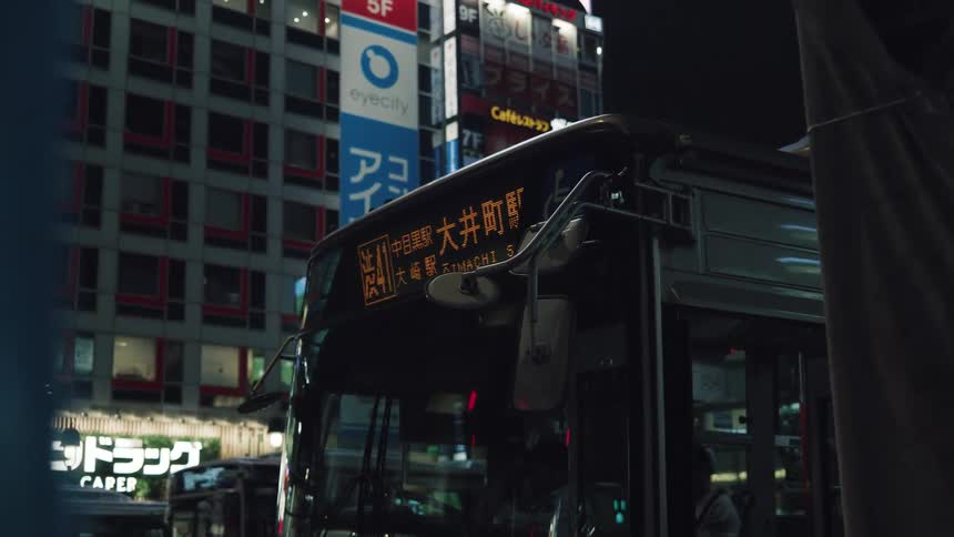 A Japanese city bus waits at a stop at night as its destination sign glows against the urban backdrop