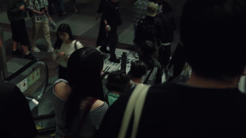 A high-angle view from behind shows commuters descending an escalator into a busy underground station at night