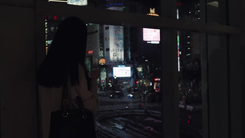 A woman stands at a window, her silhouette framed against the vibrant, bustling nightlife of a city in Tokyo