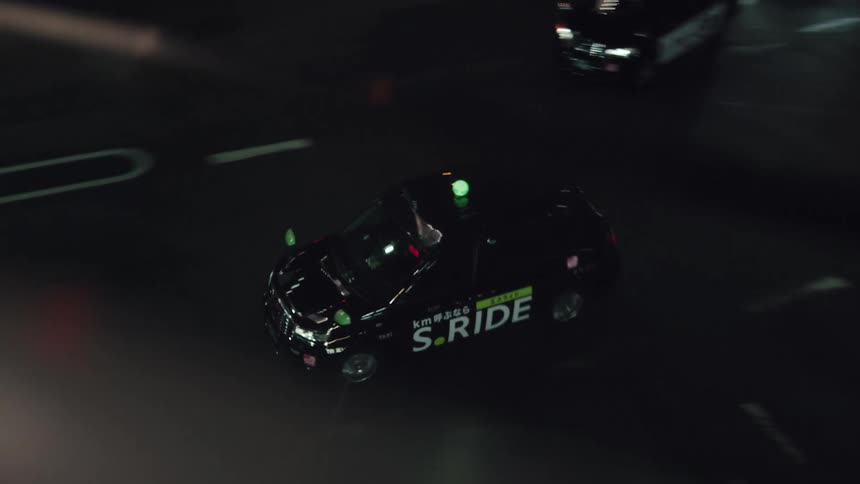 A high-angle view tracks a black taxi as it drives along a dark city street at night in Japan