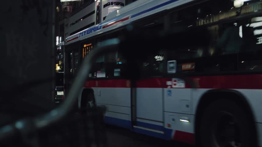 A city bus departs from a stop and drives through the neon-lit streets of Tokyo at night