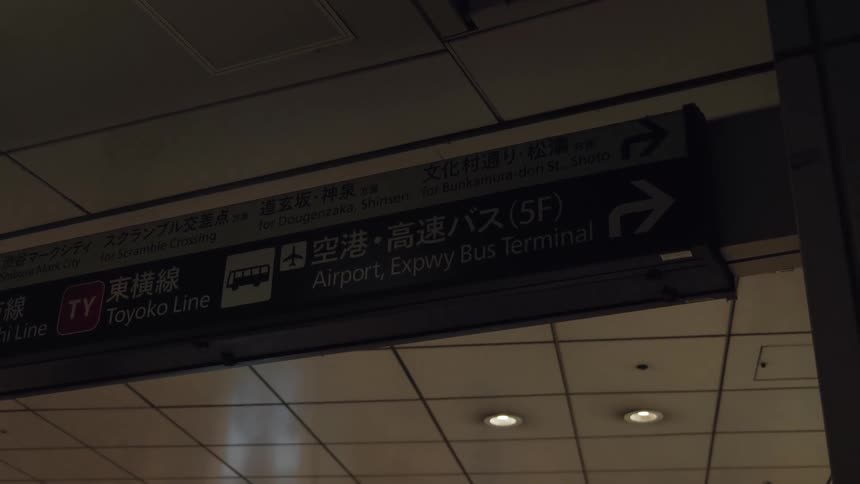 A low-angle shot of a dimly lit directional sign in a Japanese transit station pointing towards the airport and bus terminal