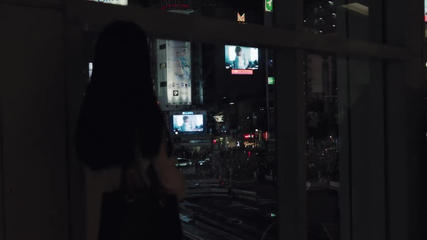 A woman with long dark hair looks out a window over the bustling, neon-lit streets of Tokyo at night