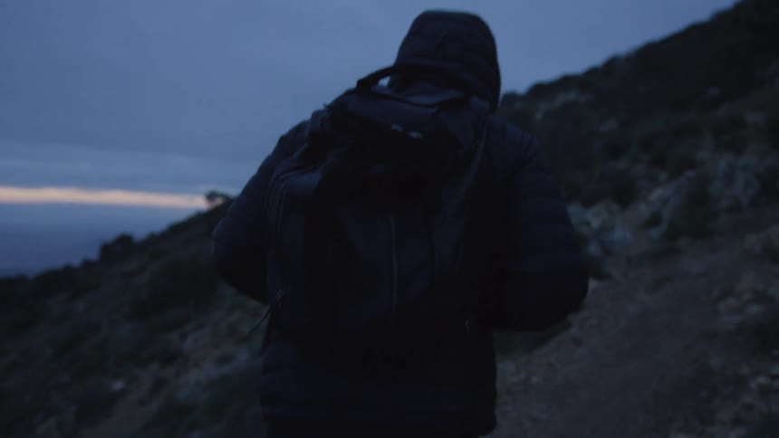 A person wearing a dark hooded jacket and a backpack hikes up a mountain trail at dusk in slow motion