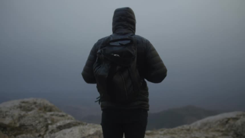 A person in a hooded jacket and backpack stands on a rocky mountain peak, looking out at the foggy landscape below
