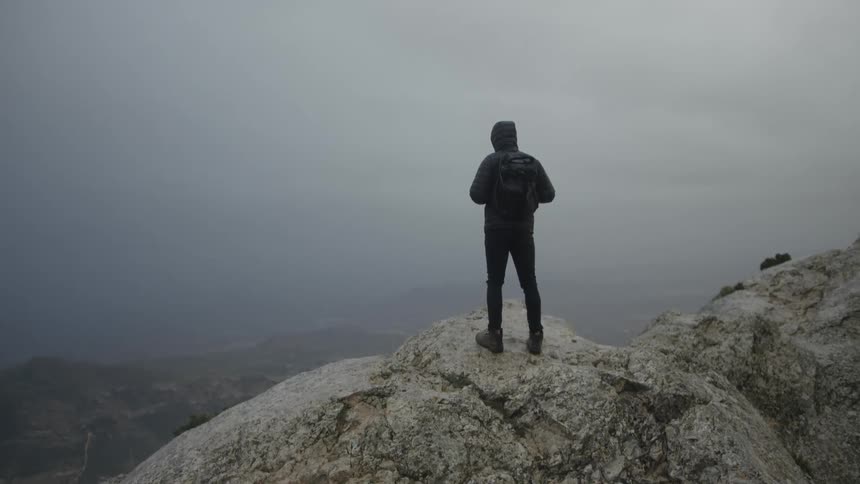 A solitary hiker in a hooded jacket stands on a rocky mountain peak, looking out at the vast, foggy landscape below on an overcast day