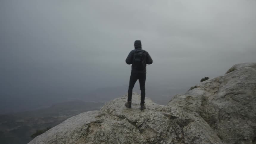 A lone hiker with a backpack stands on a rocky mountain summit, looking out over a vast, foggy landscape on an overcast day