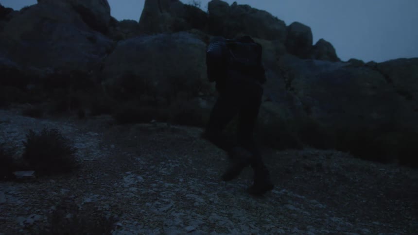 Slow motion shot of a hiker with a backpack ascending a rocky path in a dark, mountainous landscape