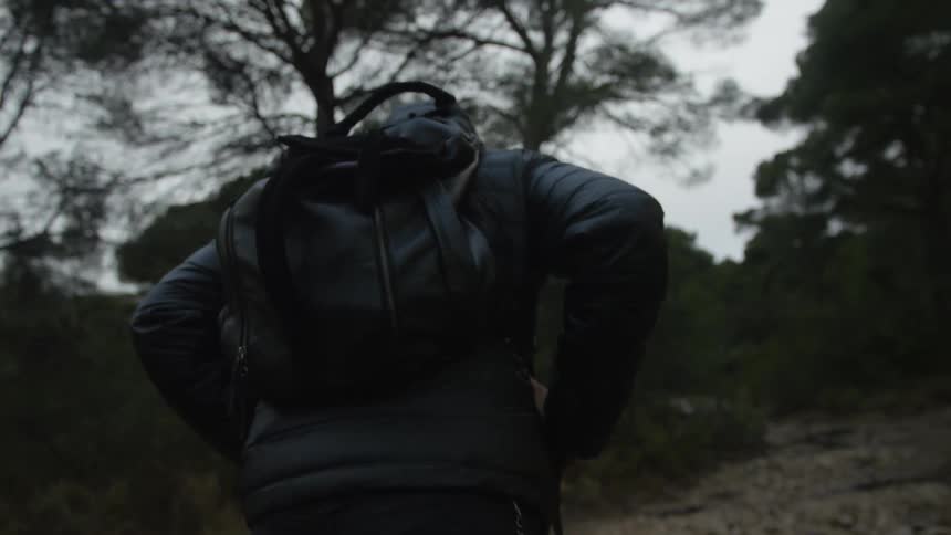 Slow motion shot from behind of a person in a black hooded jacket and backpack hiking through a dark, gloomy forest