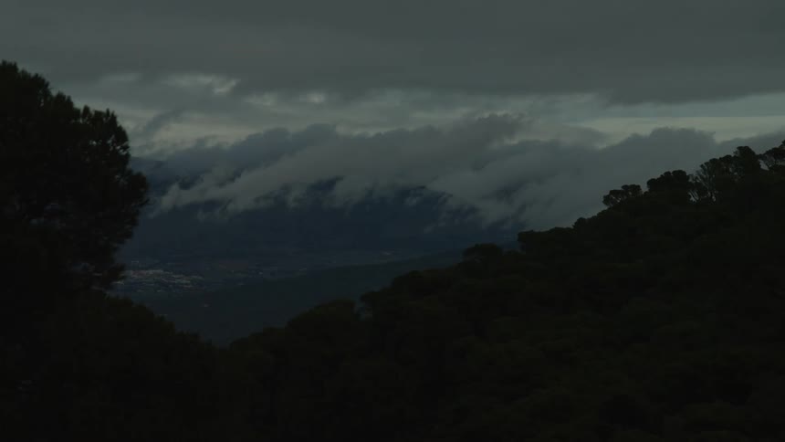 Ominous storm clouds hover over distant mountains, with a silhouetted forest in the foreground and city lights twinkling in the valley below