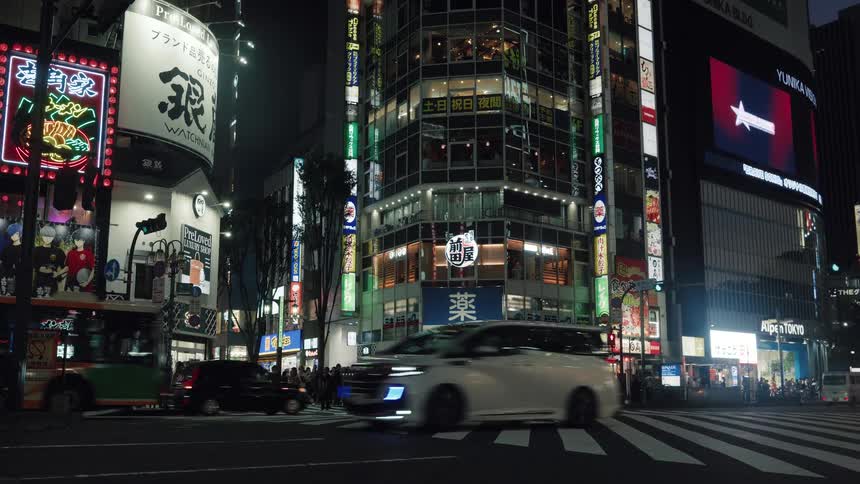 Traffic flows through a bustling Tokyo intersection at night as crowds of people wait to cross under vibrant neon signs