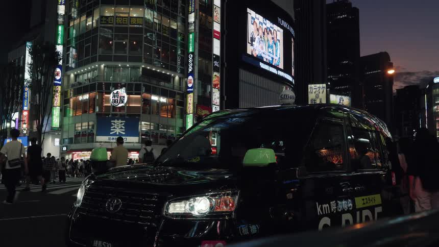 A black taxi waits at a bustling Tokyo intersection at night as pedestrians cross the street under bright neon signs