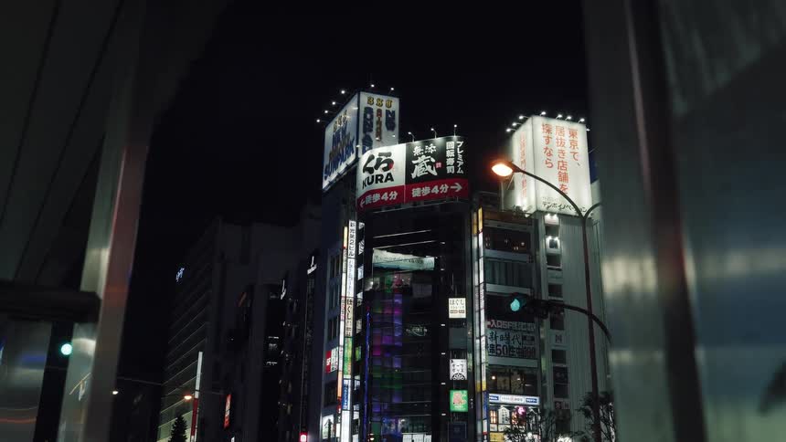 A slow-moving shot captures the towering, neon-lit skyscrapers of a Tokyo street corner at night, framed by an overpass