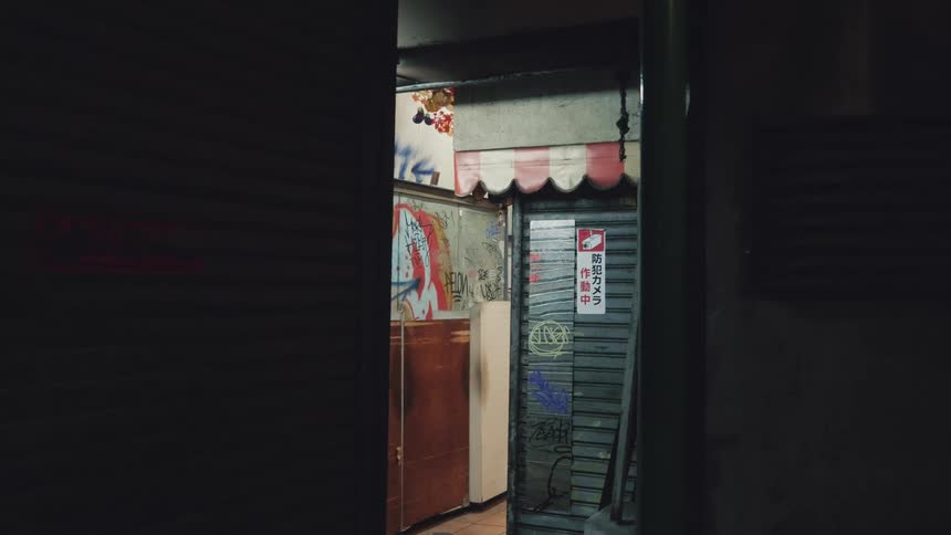 A view into a dark and gritty urban alleyway at night reveals a graffiti-covered storefront with a Japanese security sign