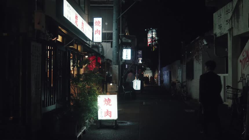 A man walks away from the camera down a dark alley in a Japanese city at night, illuminated by glowing restaurant signs