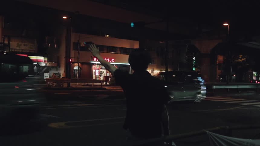 A person waves to passing cars at a busy intersection in a Japanese city at night