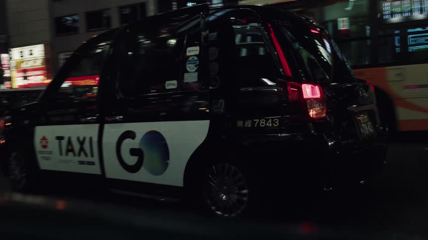 A Japanese taxi cab with a passenger inside automatically closes its door and drives off into the brightly lit city streets of Tokyo at night
