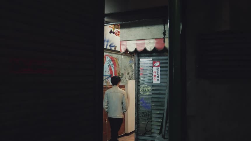 A man walks down a dark, graffiti-covered alleyway between closed storefronts in a city at night