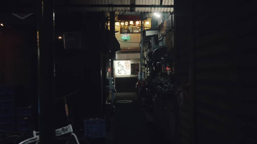 A dark, narrow alleyway in Tokyo leads to a brightly lit restaurant with glowing signs at night