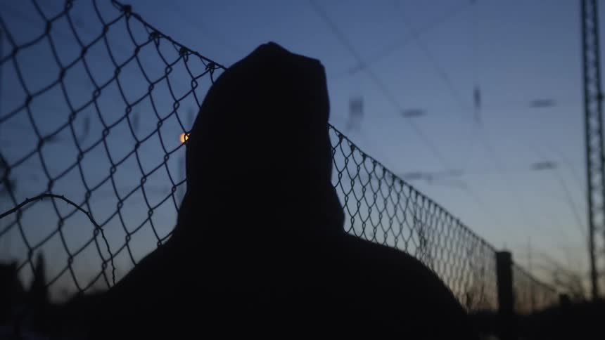 A lonely figure in a hoodie is silhouetted against the twilight sky as they look through a chain-link fence
