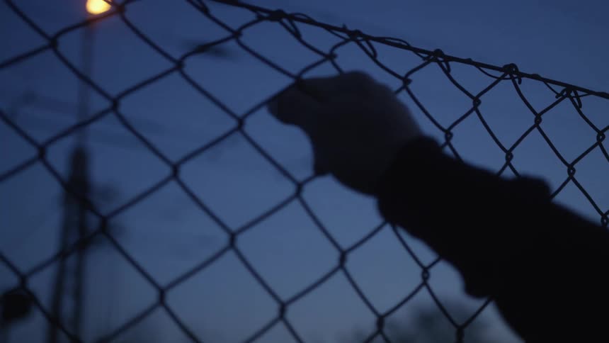 A silhouetted hand slowly reaches for and grips a chain-link fence topped with barbed wire against a dark evening sky