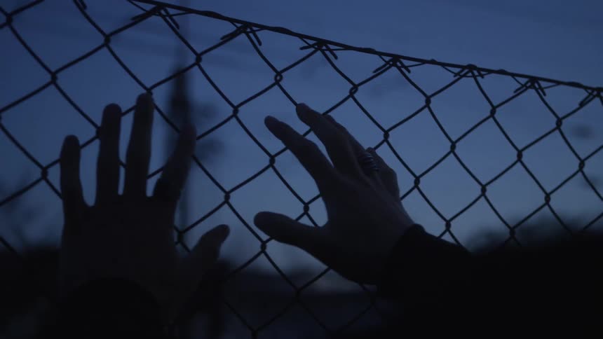 Silhouetted hands grip a chain-link fence at dusk, conveying a sense of confinement and desperation